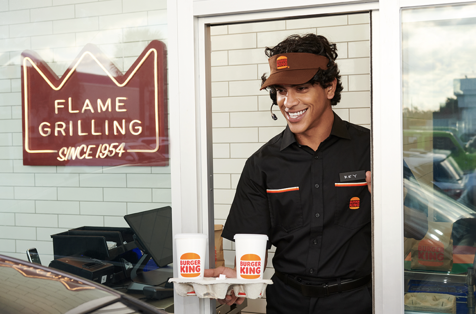 Burger King drive-thru team member holding two branded drink cups beside the Flame Grilling neon sign