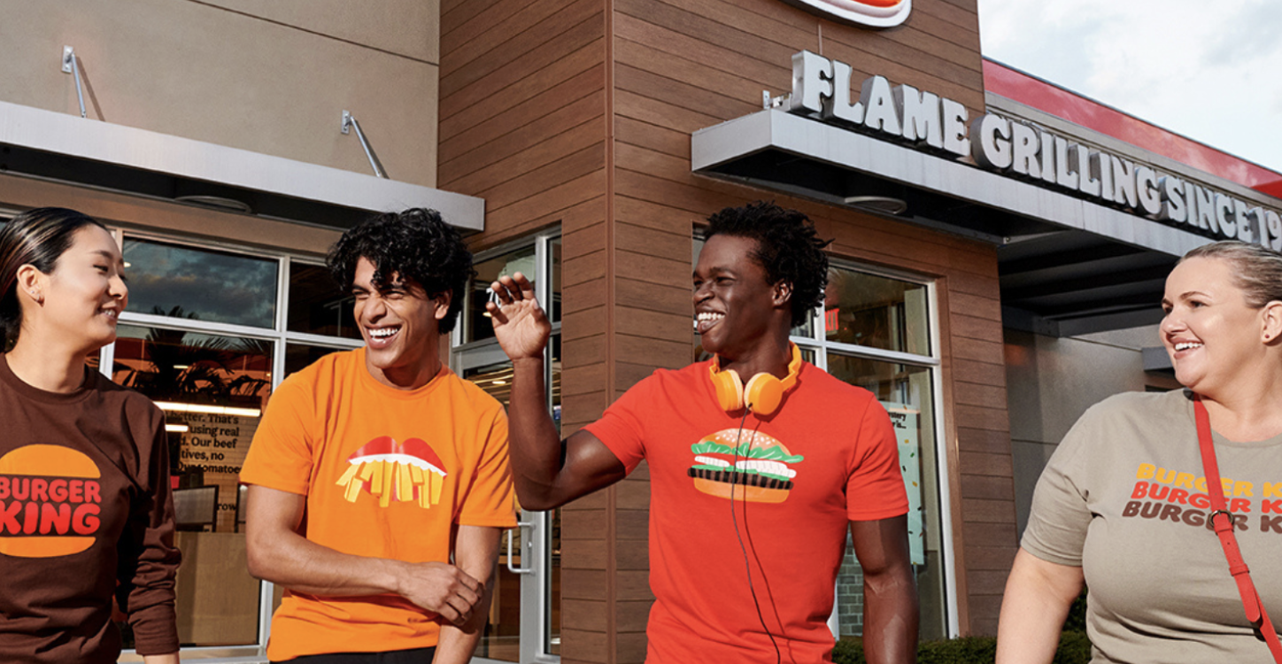 Four smiling Burger King team members in branded tees walking together outside a Flame Grilling restaurant
