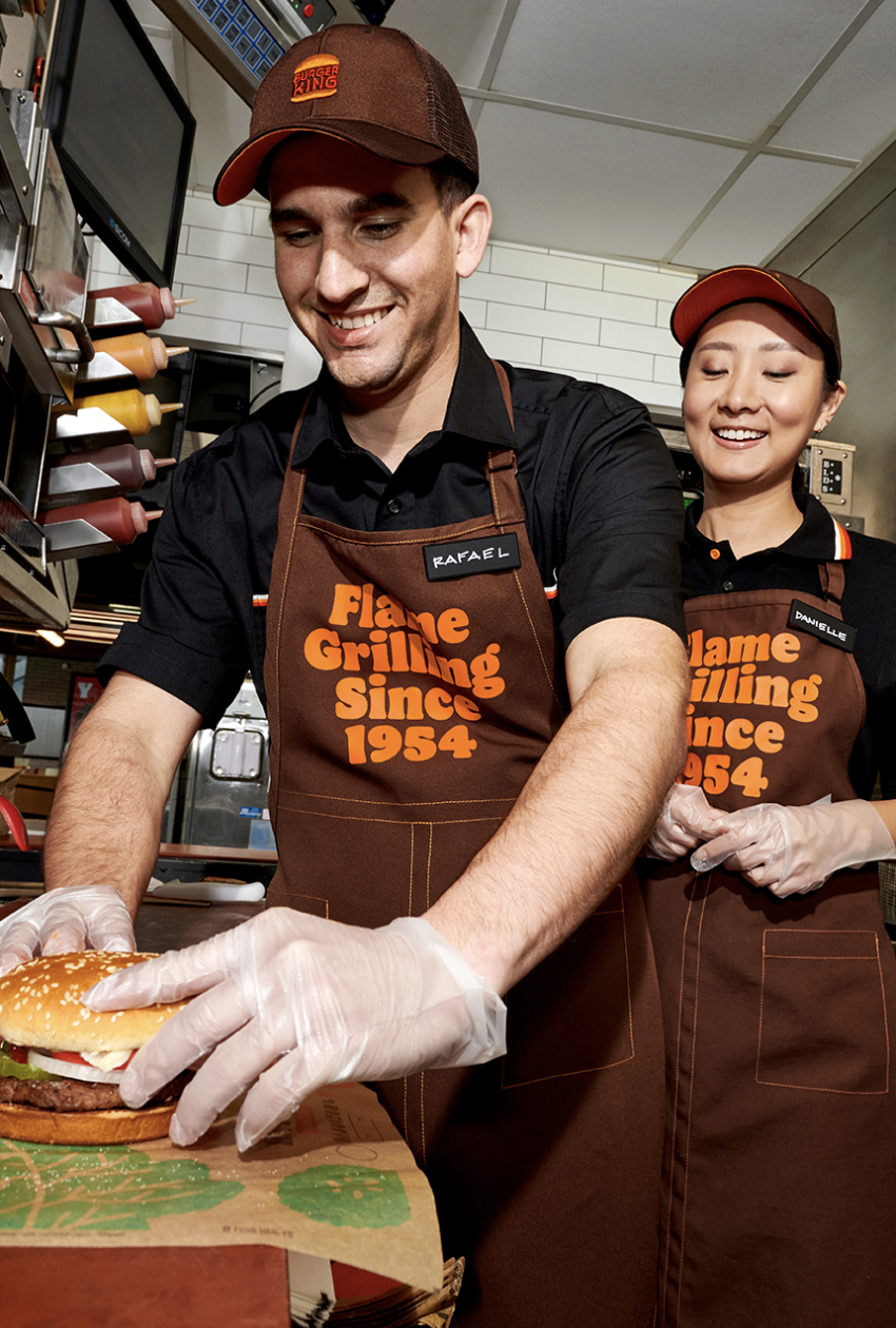 Two Burger King team members in Flame Grilling aprons plating a freshly grilled burger in the kitchen