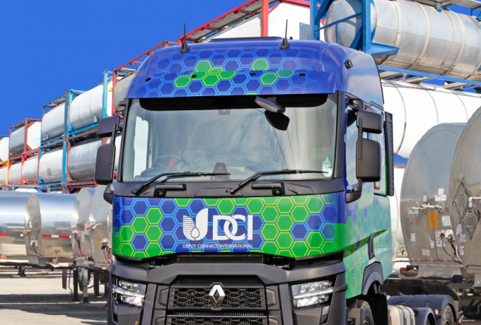 A blue and green DCI tank truck parked among rows of stainless steel ISO tank containers in a depot yard