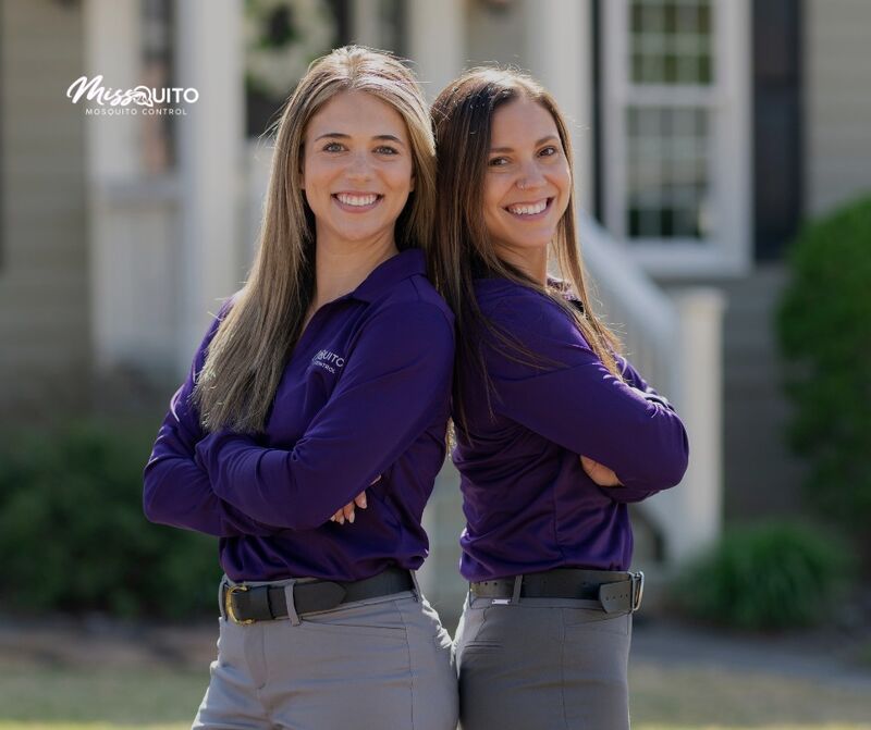 Two MissQuito team members in purple uniforms smiling together in front of a home