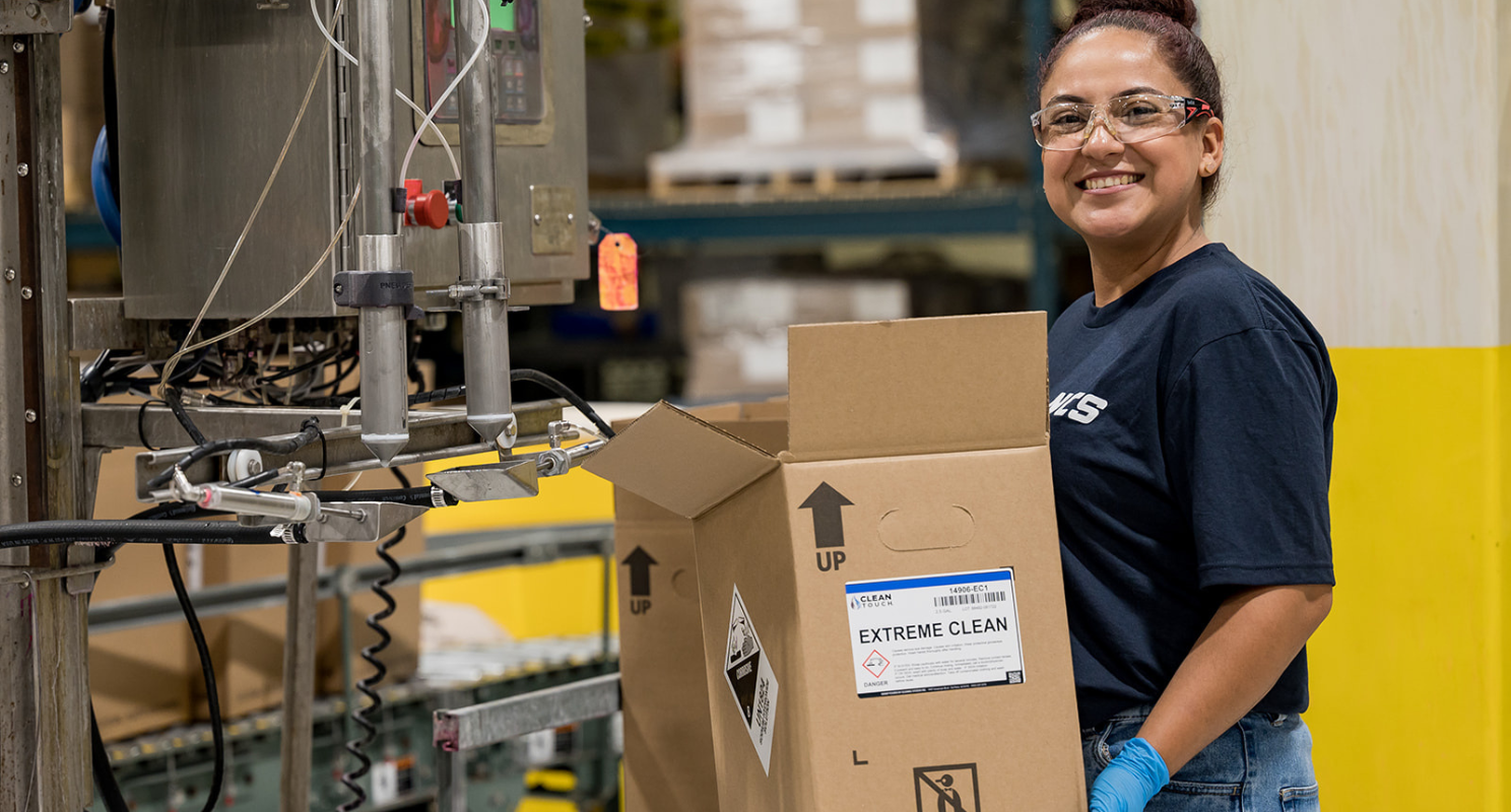 An NCS team member in a branded shirt and safety glasses packaging Extreme Clean carwash chemistry on the production floor