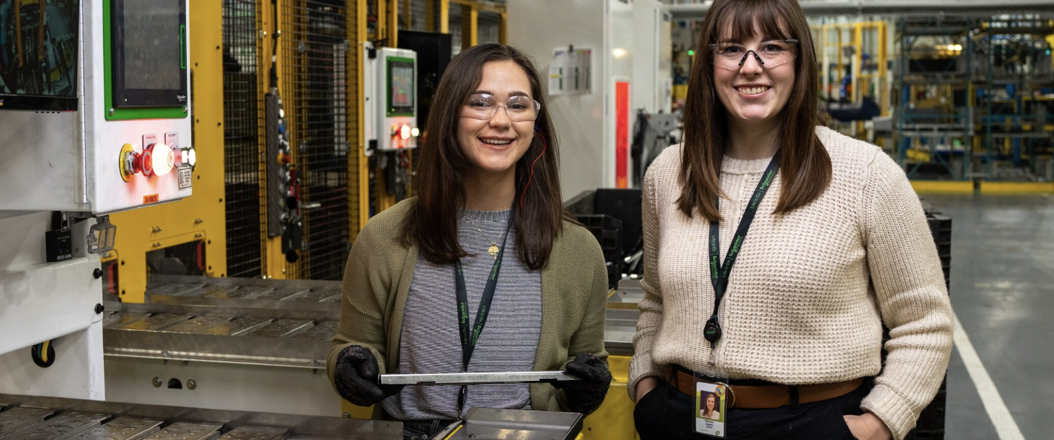 Two Schneider Electric employees in safety glasses beside automation control equipment on a factory floor