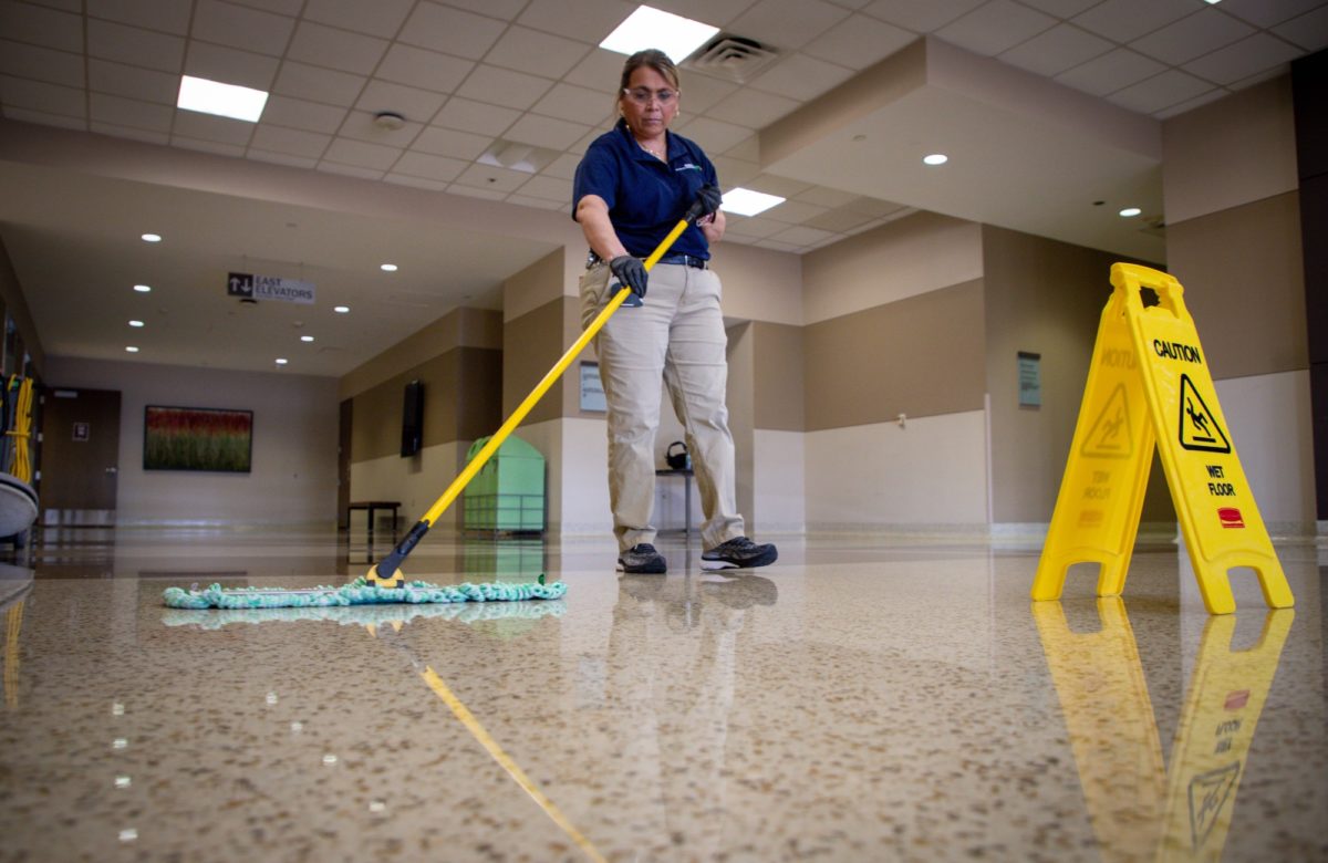 An SSC custodian in a school building mopping a polished floor beside a wet floor caution sign