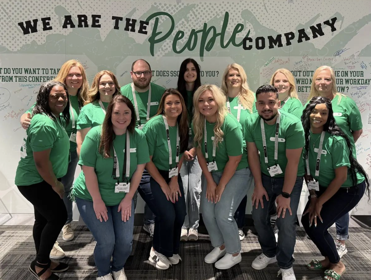 SSC team members in green shirts smiling together in front of a "We Are The People Company" wall at a company event