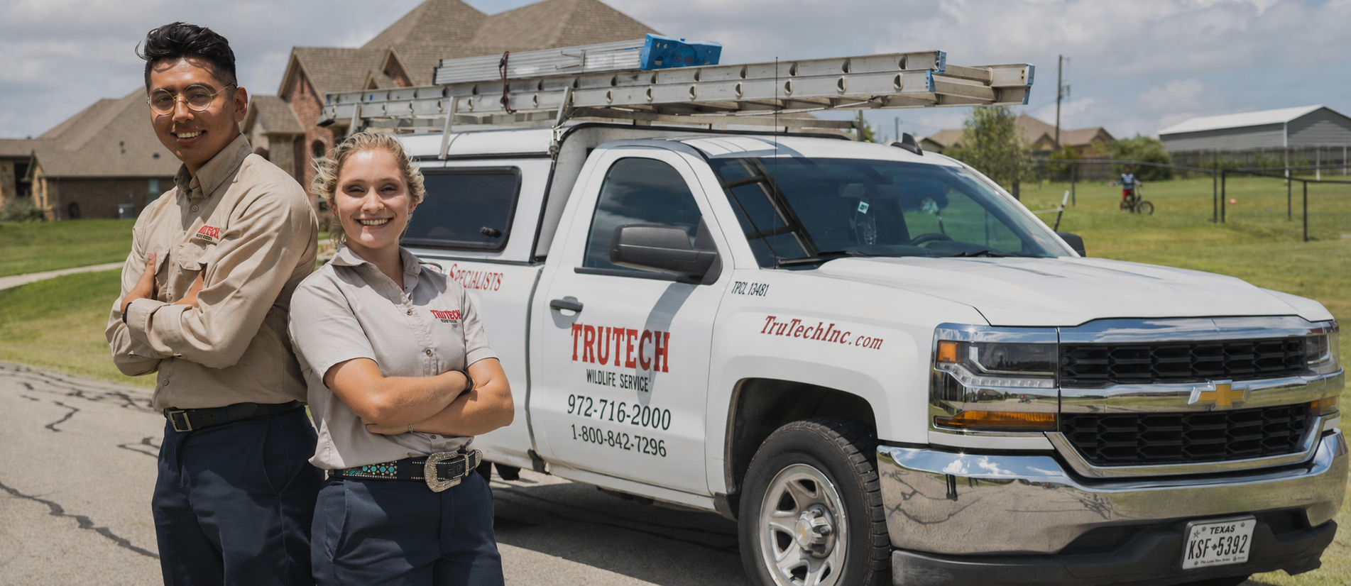 Two smiling Trutech Wildlife Service technicians standing beside a service truck in a residential neighborhood