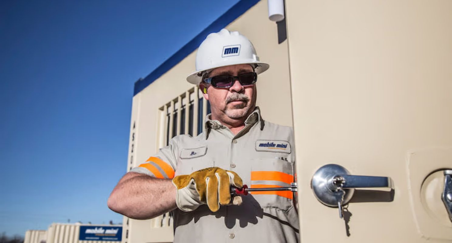 WillScot Mobile Mini technician in a hard hat and hi-vis vest using a tool to service a portable storage container