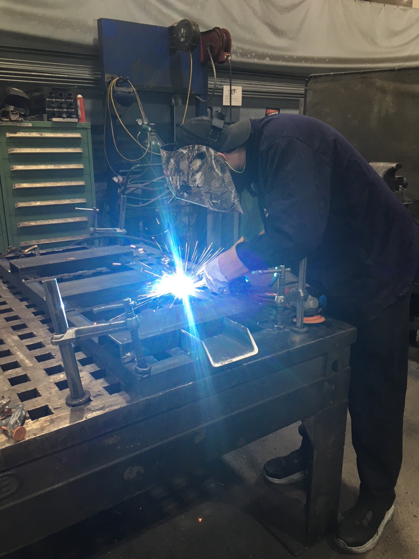 A man welding metal in a Harley-Davidson workshop, focused on his task with protective gear and tools surrounding him